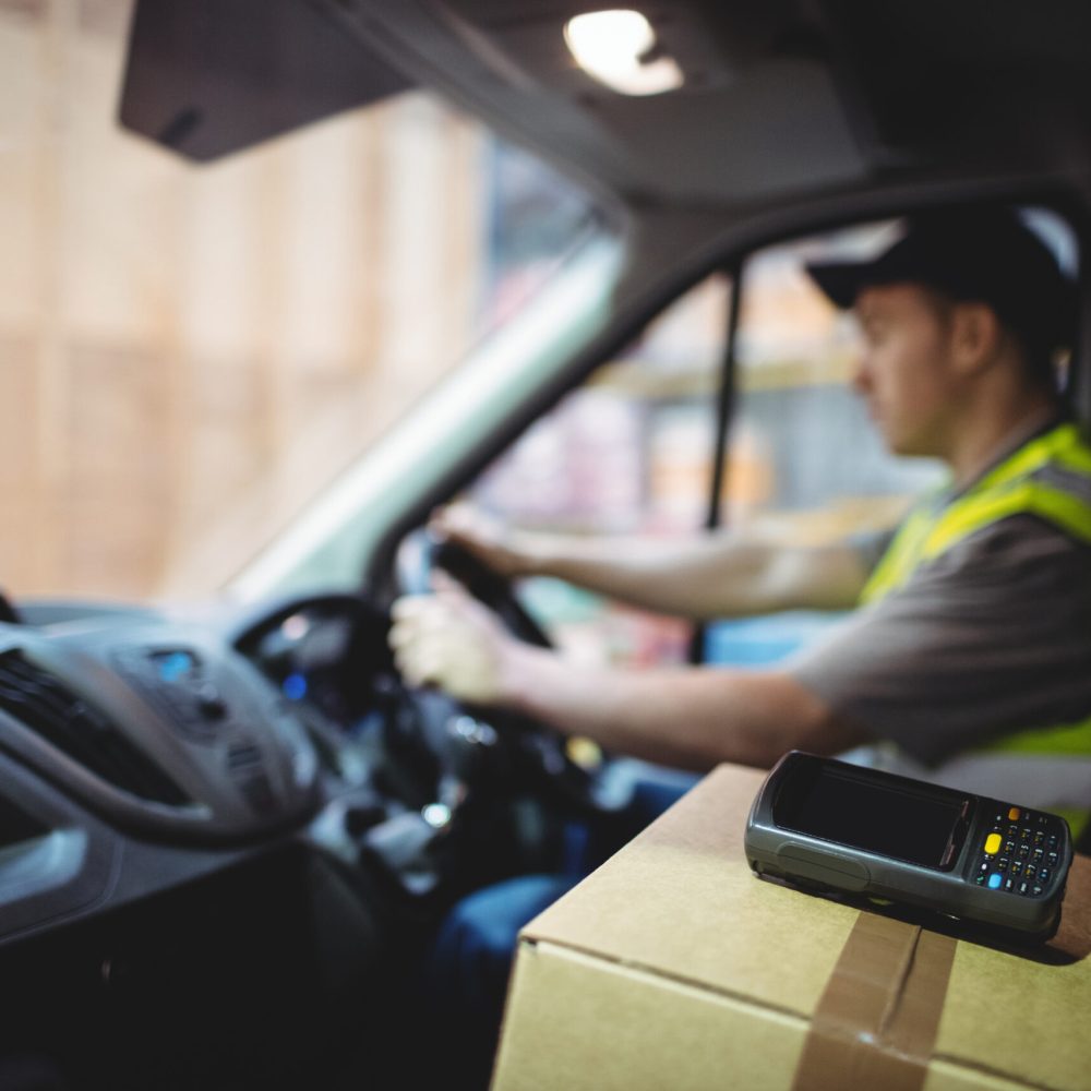 Delivery driver driving van with parcels on seat outside warehouse